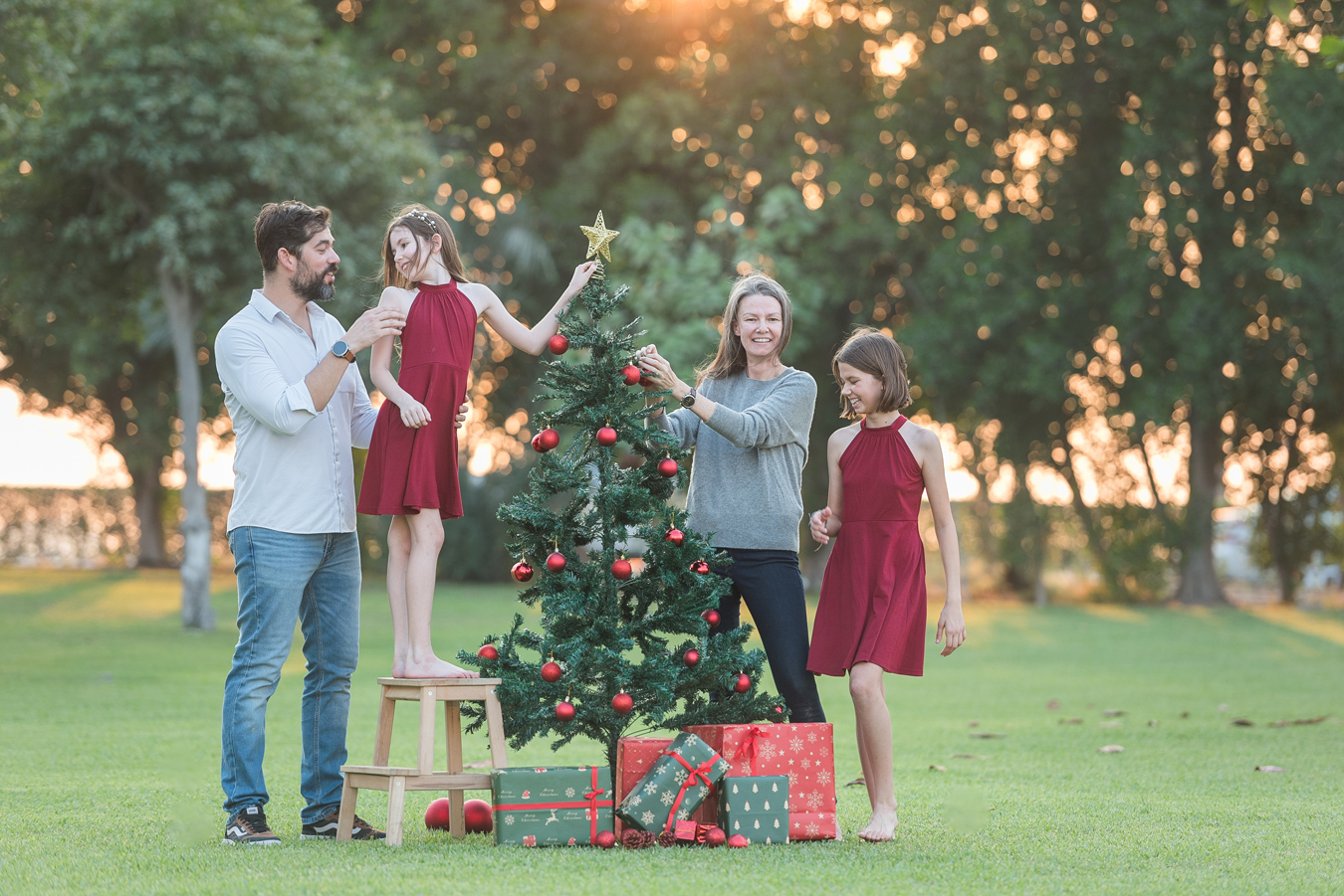 Family decorating outdoor Christmas tree with red ornaments and gifts. Two girls in red dresses help parents